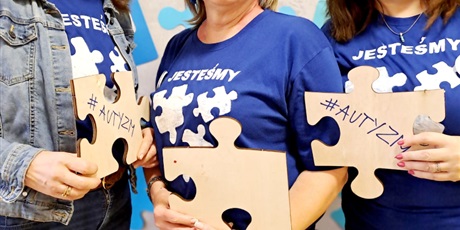 Powiększ grafikę: Nauczycielki z drewnianymi puzzlami w ręku i niebieskimi koszulkami z namalowanymi białymi puzzlami pozują do zdjęcia, w tle mata z niebieskimi puzzlami na kremowym tle