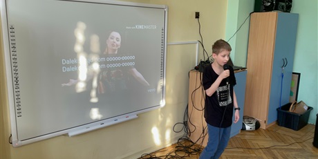 Powiększ grafikę: Chłopiec stoi z mikrofonem i śpiewa, Za nim znajduję się monitor z tekstem piosenki