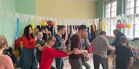 Powiększ grafikę: Wychowankowie z nauczycielami tańczą w kółku. Sala jest udekorowana białą bibułą i kolorowymi balonami