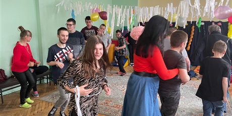 Powiększ grafikę: Wychowankowie z nauczycielami tańczą w kółku. Sala jest udekorowana białą bibułą i kolorowymi balonami