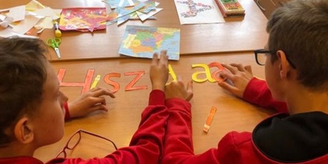 Powiększ grafikę: 3.	uczniowie układają na ławce napis Hiszpania z kolorowych, żółto-czerwonych liter. W tle, na ławce, leżą obrazki herbu i mapy Hiszpanii.