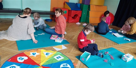 Powiększ grafikę: uczniowie i nauczyciele siedzą na podłodze grając w duże gry planszowe