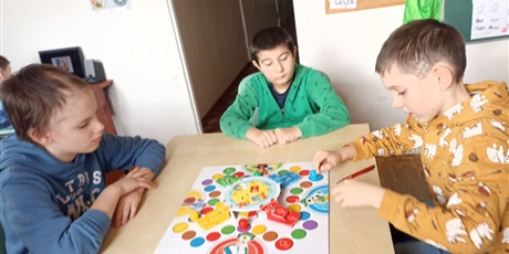 Powiększ grafikę: Uczniowie grają w grę planszową, w tle tablica szkolna