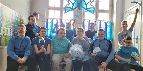 Powiększ grafikę: Grupa chłopców i dziewczynek ubrana na niebiesko  siedzi na krzesłach, w rękach trzymają niebieskie  papierowe motyle.