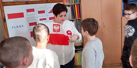 Powiększ grafikę: nauczycielka pokazuje uczniom jak wygląda flaga Polski. Dzieci ułożą potem flagę z pasów w odpowiednim kolorze