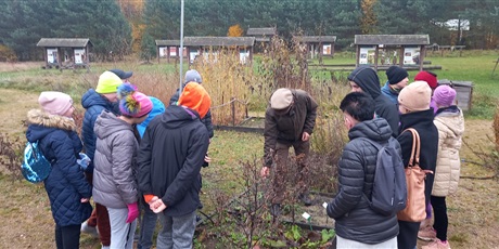 Powiększ grafikę: uczniowie stoją wokół rabaty i słuchają leśniczego, który pokazuje rośliny