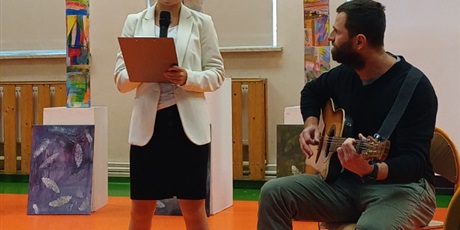 Powiększ grafikę: Uczennica śpiewa piosenkę na akademii, obok nauczyciel gra na gitarze, w tle dekoracje.