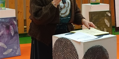 Powiększ grafikę: Nauczyciel stoi z mikrofonem i czyta z kartki położonej na kubiku. W tle dekoracje.