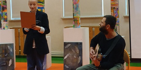 Powiększ grafikę: Uczeń śpiewa piosenkę na akademii, obok nauczyciel gra na gitarze, w tle dekoracje.