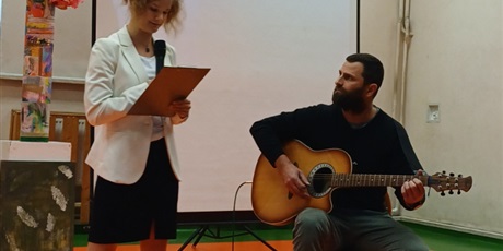 Powiększ grafikę: Uczennica śpiewa piosenkę na akademii, obok nauczyciel gra na gitarze, w tle dekoracje.