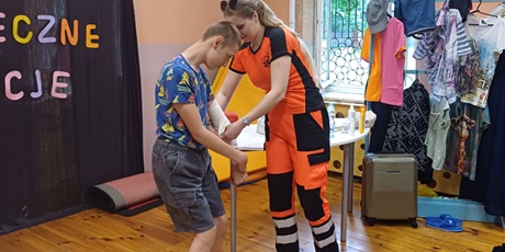 Powiększ grafikę: Pani ratownik bandażuje rękę chłopcu. W tle wieszak z ubraniami i walizka.
