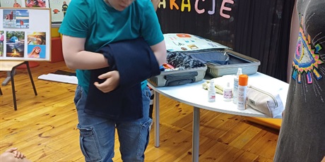 Powiększ grafikę: Chłopiec składa spodnie. Na stole otwarta walizka z popskładanymi ubraniami. Na stole wakacyjne kosmetyki. w tle napis " Bezpieczne wakacje"
