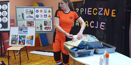 Powiększ grafikę: Pani ratowniczka  zamyka zapakowaną ubraniami walizkę leżącą na stole. W tle tablice demontracyjne.