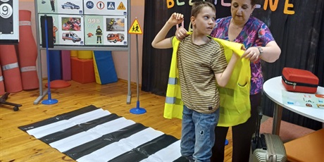 Powiększ grafikę: Nauczycielka zakłada chłopcu na plecy kamizelkę odblaskową. Obok leżą na podłodze pasy  uliczne i stoi tablica demonstracyjna z numerami alarmowymi.