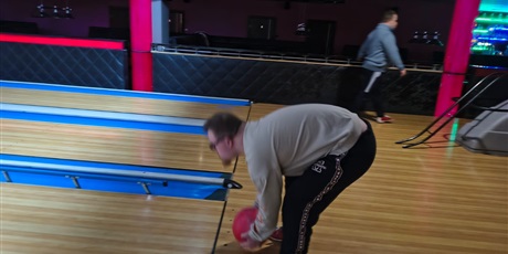 Powiększ grafikę: Widok z boku: dziecko oburącz rzuca kulą po torze, w tle inny zawodnik.