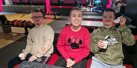 Powiększ grafikę: Uśmiechnięci zawodnicy siedzą przystole w kręgielni, w tle tory i kule.