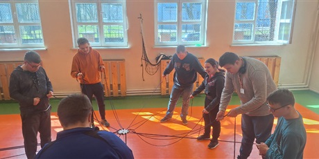 Powiększ grafikę: Uczniowie stoją w kółku trzymając sznurki za pomocą których wspólnie ustawią wieżę z klocków. W tle okna sali gimnastycznej.