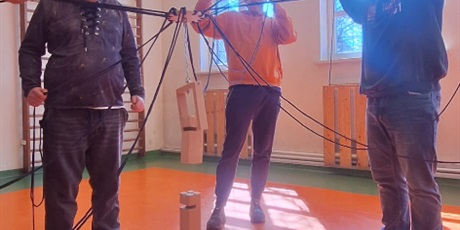 Powiększ grafikę: Uczniowie stoją w kółku trzymając sznurki ustawili wieżę z klocków. W tle okna sali gimnastycznej.