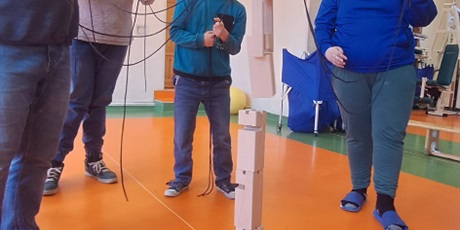 Powiększ grafikę: Uczniowie stoją w kółku trzymając sznurki za pomocą których wspólnie ustawią wieżę z klocków. W tle przyrządy sportkowe.