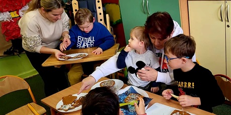 Powiększ grafikę: Jeden przedszkolak dekoruje pierniczki z wychowawczynią drugi chłopiec robi to samo wspólnie z rodziną. W tle widać choinkę i plakat świętego Mikołaja