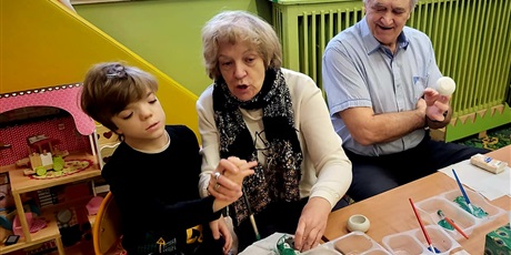 Powiększ grafikę: Babcia i dziadek malują z wnukiem świeczniki. W tle meble.