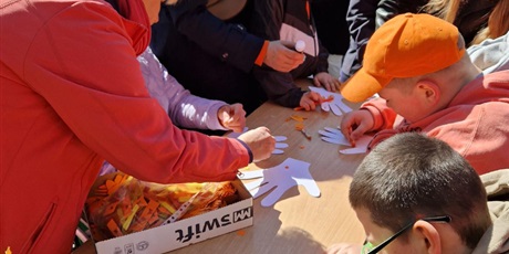 Powiększ grafikę: Nauczycielka z dziećmi tworzy pomarańczowe dekoracje na szablonach dłoni rozłożonych na stole.