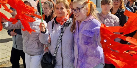 Powiększ grafikę: Nauczycielka trzma z dłoni pomarańczową ozdobę, obokstoi uczennica. W tle pomarańczowy tłum.