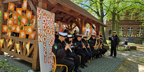 Powiększ grafikę: Koncert orkiestry, która siedzi w udekorowanej na pomarańczowo wiacie z drewana. W tle budynek z cegły.