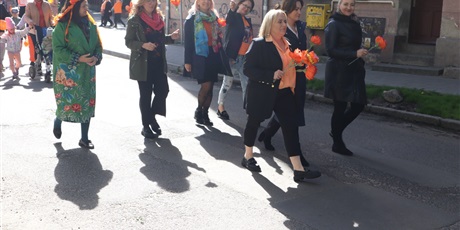 Powiększ grafikę: 7 kobiet kolorowo ubranych, trzymających w rękach pomarańczowe rzeczy idą w pochodzie, za nimi, w tle wychowawczyni przedszkola z przedszkolakami.