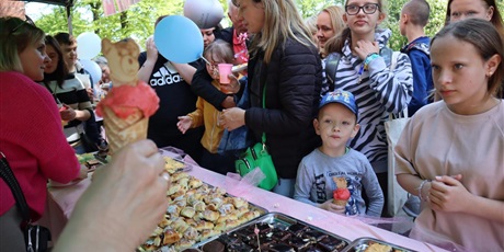 Powiększ grafikę: Dzieci z rodzicami w kolejce po lody i ciasta, w tle zieleń drzew