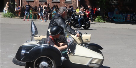 Powiększ grafikę: Motocykl z koszem pasażerskim , mężczyzna przewozi dziecko w kasku, w tle inny motocykl, grupa ludzi i budynek.