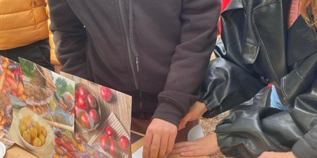 Powiększ grafikę: Chłopiec i wychowawczyni stoją przy stole z prezentacją zdjęć dotyczących ziemniaków. Chłopiec odciska na papierze żółtą pieczątkę zrobioną z ziemniaka.