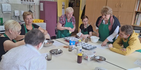 Powiększ grafikę:  Kobiety i młodzież wokół stołu, na którym stoją naczynia, przybory kuchenne oraz produkty spożywcze trzymają produkty do wykonania ciasta. Na stole widoczne są foremki do babeczek.