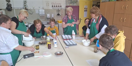 Powiększ grafikę: Kobiety i młodzież wokół stołu, na którym stoją naczynia, przybory kuchenne oraz produkty spożywcze przygotowują ciasto, mieszają produkty, miksują mikserem ręcznym.