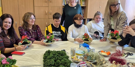 Powiększ grafikę: Osoby siedzą przy stole, trzymają przed sobą naczynia z kwiatami. Na środku stołu rozłożone  dekoracje.
