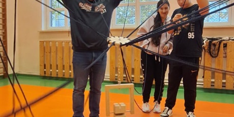 Powiększ grafikę: Dwóch uczniów i uczennica trzymają liny z kwiatem zawieszonym nad klockami drewnianymi. W tle okna sali gmnastycznej.