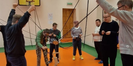 Powiększ grafikę: Siedmioro uczniów trzyma liny z kwiatem zawieszonym nad klockami drewnianymi. W tle ściany i drzwi sali gmnastycznej.