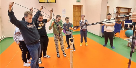 Powiększ grafikę: Siedmioro uczniów trzyma w oddali liny z kwiatem zawieszonym nad klockami drewnianymi. W tle ściany i drzwi sali gimnastycznej.