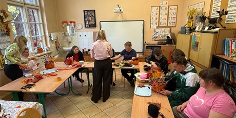 Powiększ grafikę:  Stoły są ustawione  w pól kregu przy nich siedzą uczniowie, którzy wraz z   nauczycielami przystrajaja dynie: liscmi, włóczkami, kasztanami.