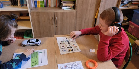 Powiększ grafikę: Dwóch uczniów siedzi przy stole i pracują na zalaminowanych kartach pracy.Jeden dopasowuje litery pod obrazkiem miotły. Drugi w słuchawkach do pasowuje symbole do obrazków w temacie sprzątacze.
