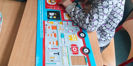 Powiększ grafikę: Chłopiec siedzi przy dużych puzzlach wozu strażackiego i do okrągłych okienek wciska brakujące elementy wyposażenia strażackiego.