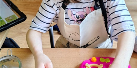Powiększ grafikę: Dziewczynka w faruszku siedzi i bawi się przy stole akcesoriami kucharskimi. Zdejmuje z patelni zabawkowe jajko sadzone.