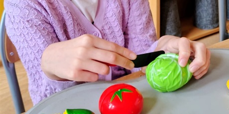 Powiększ grafikę: Dziewczynka siedzi przy stole. Na podkładce ma plastikowy zestaw owoców i warzyw do przekrajania na pół. Kroi nożem.