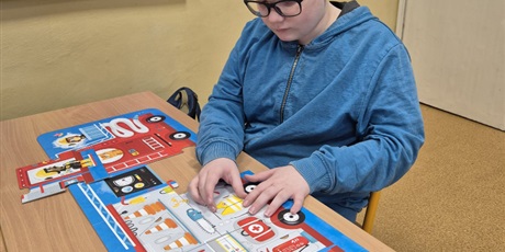 Powiększ grafikę: Chłopiec układa przy stoliku duże puzzle wóz strażacki.