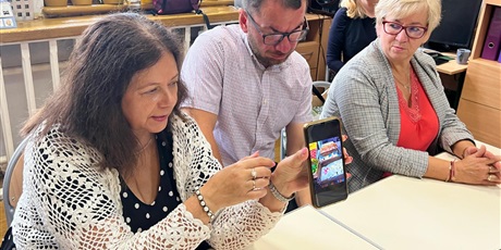 Powiększ grafikę: Dwie kobiety, pomiędzy nimi mężczyzna siedzą przy biurku, kobieta z lewej demonstruje telefon komórkowy, w tle okna.