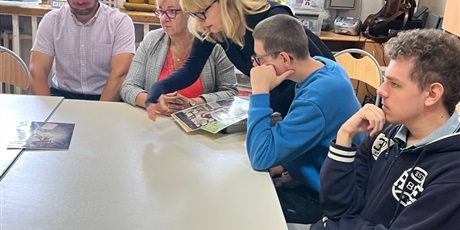 Powiększ grafikę: Mężczyzna, dwie kobiety i dwoje uczniów siedzą za biurkiem i przeglądają czasopismo, w tle okna