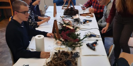 Powiększ grafikę: uczniowie wraz z rodzicami, opiekunami siedzą przy wspólnym stole i wybierają elementy do produkcji stroików świątecznych