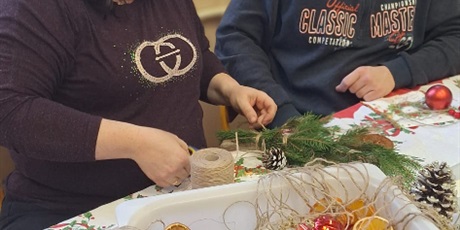 Powiększ grafikę: uczeń z mamą tworzą stroik świąteczny z gałęzi świerkowych, bombek, patyków, szyszek i kolorowych koralików