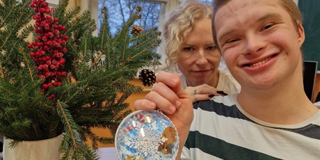 Powiększ grafikę: uczeń z mamą prezentują wykonaną wspólnie bombkę na tle stroika z gałęzi świerkowych