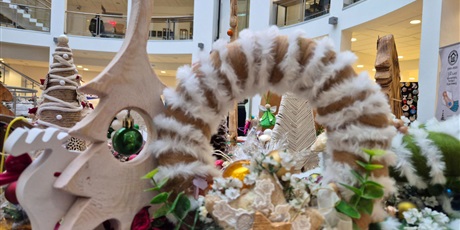 Powiększ grafikę: wianek owinięty białą włóczką o bardzo miękkiej i puszystej fakturze ozdobiony gwiazdą, sztucznymi kwiatami i bombką. Obok wianka stoi mała, biała, drewniana choinka ozdobiona zieloną bombką i gipsowy